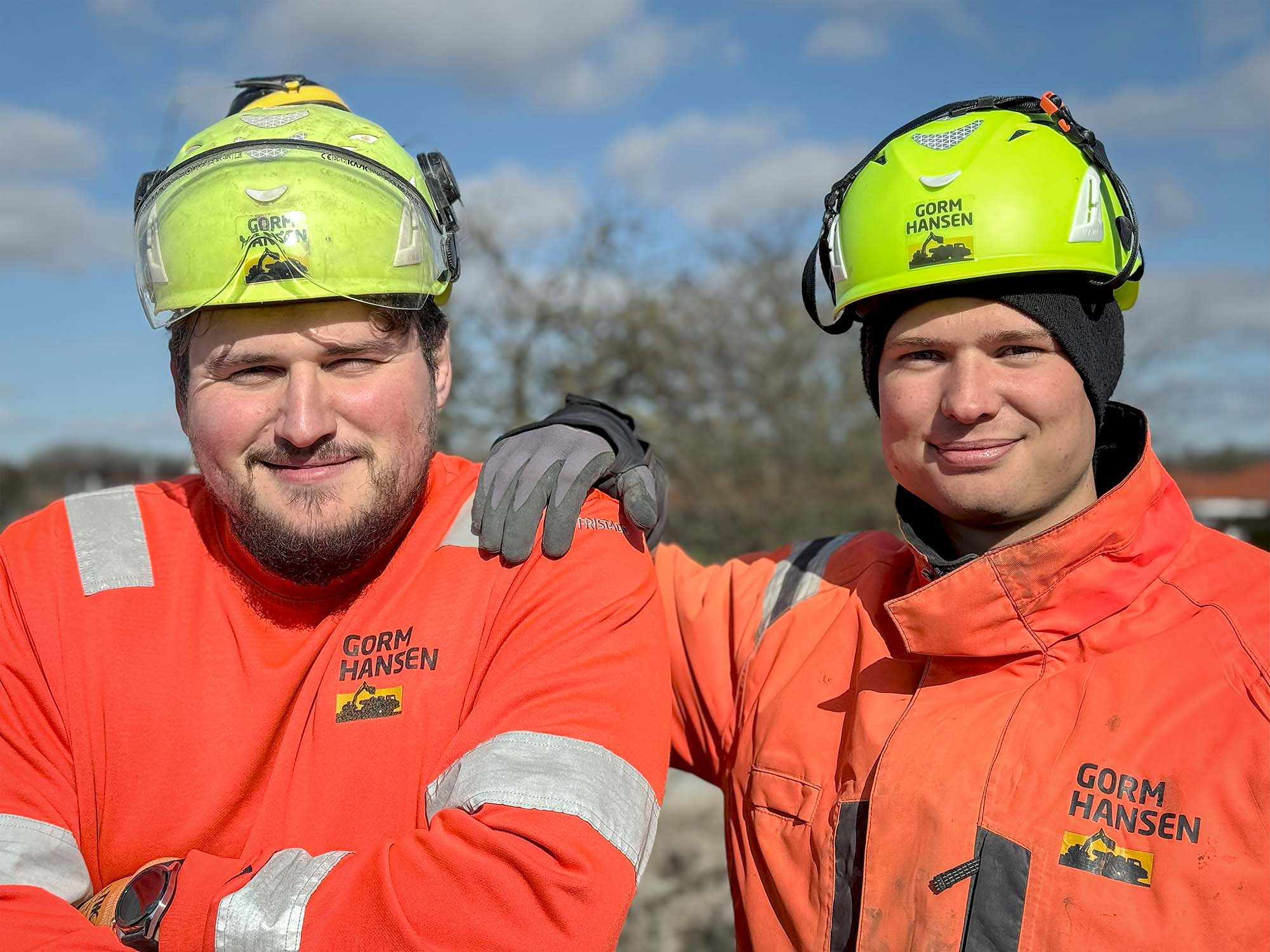 To bygningsarbejdere, iklædt orange sikkerhedstøj med reflekterende striber og neongule hjelme med “Gorm Hansen”-logoet, står tæt sammen udendørs. Den ene har armen om den anden, begge smilende under en overskyet himmel. To bygningsarbejdere, iklædt orange sikkerhedstøj med reflekterende striber og neongule hjelme med "Gorm Hansen"-logoet, står tæt sammen udendørs. Den ene har armen om den anden, begge smilende under en overskyet himmel.